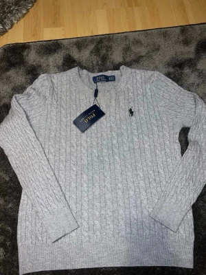 Ljusgrå kabelstickad tröja Polo Ralph Lauren - Snygg grå kabelstickad tröja från Polo Ralph Lauren med klassisk rund halsringning och broderad logga på bröstet. Tröjan har långa ärmar och ribbade muddar vid ärmslut och nederkant. Perfekt för en stilren och avslappnad look.                   Bilderna är ej tagna av mig, från förra ägaren men är ej använd sen jag köpte den                                       Kan vara för tight om du är över 170, är 178 och passar inte mig 