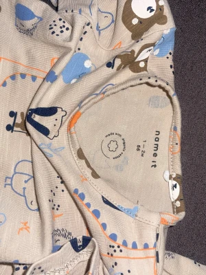 Beige body och byxa med djurmönster - Sött set från Name It med långärmad beige body och matchande byxa. Båda plaggen har gulligt djurmönster i blått, orange och brunt med björnar, fåglar och andra djur. Tillverkad i mjuk bomull, perfekt för små barn.