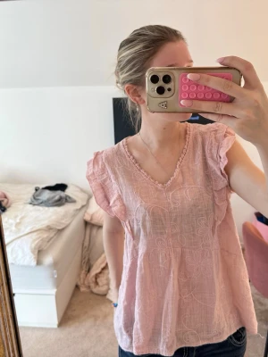 Ljusrosa blus med broderi och volang - Söt ljusrosa blus med v-ringning, broderade detaljer och volang på ärmarna. Blusen är luftig och har ett lätt transparent material som ger en avslappnad känsla. Perfekt att matcha med jeans eller kjol.