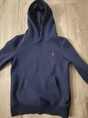 Mörkblå hoodie från Polo Ralph Lauren - Snygg mörkblå hoodie från Polo Ralph Lauren i slim fit-modell. Klassisk huva, känguruficka framtill och den ikoniska röda loggan broderad på bröstet. Perfekt för en avslappnad och stilren look. Tillverkad i mjukt bomullsmaterial. Nypris 1000 kr.