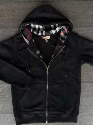 Svart hoodie från Burberry - Svart hoodie från Burberry med hel dragkedja och klassiskt rutigt foder i huvan. Tröjan har två fickor framtill, ribbade muddar och snörning i huvan. Perfekt för en stilren och avslappnad look.