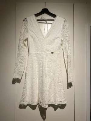Vit spetsklänning med v-ringning - Kort vit spetsklänning med lång ärm och djup v-ringning både fram och bak. Klänningen har ett omlottliknande liv och knytband i nacken. Tillverkad i spetsmaterial som ger en romantisk känsla.