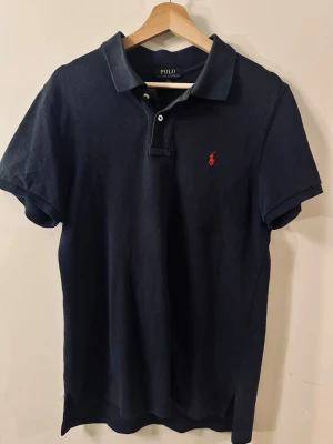 Marinblå pikétröja från Polo Ralph Lauren - Klassisk marinblå pikétröja från Polo Ralph Lauren med korta ärmar och röd broderad logga på bröstet. Tröjan har krage, knappar vid halsen och är tillverkad i bomull. Perfekt för en stilren och avslappnad look.