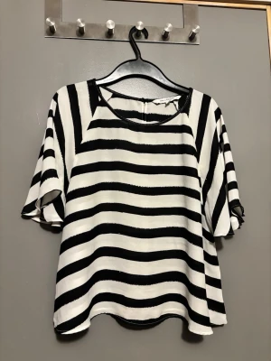 Randig svartvit blus med vida ärmar KappAhl - Säljer en loose fit blus med breda svarta och vita ränder från KappAhl. Blusen har rund halsringning och korta, vida ärmar. Materialet känns lätt och luftigt, perfekt för en avslappnad stil. 