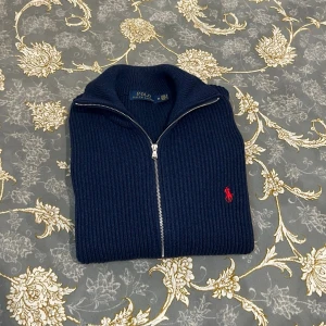 Marinblå ziptröja från Polo Ralph Lauren - Snygg marinblå stickad ziptröja från Polo Ralph Lauren med ribbad struktur och röd broderad logga på bröstet. Tröjan har hög krage, dragkedja framtill och breda muddar vid ärmslut och nederkant. Perfekt för lager på lager och stilren vardagslook.