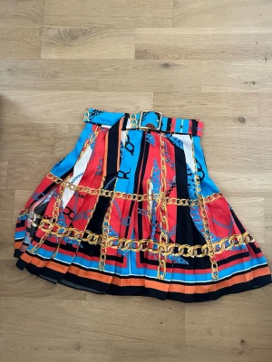 Färgglad kjol med kedjemönster från Zara - Säljer en färgstark kortkjol från Zara med bredt bälte och kedjemönster i guld. Kjolen har inslag av rött, blått, svart och orange samt plisserad design. Perfekt för dig som gillar statement-plagg och vill sticka ut.