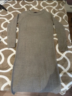 Beige stickad långklänning - Mysig beige stickad långklänning med rund halsringning och långa ärmar. Klänningen har en enkel, rak passform och ribbade muddar vid ärmslut och hals. Perfekt för kyligare dagar och enkel att styla med accessoarer.