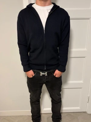 Svart hoodie med dragkedja - Säljer en svart hoodie med dragkedja framtill och huva. Hoodien är ribbstickad och har en enkel, stilren design utan synliga loggor eller tryck. Passar perfekt till en avslappnad streetwear-look och är lätt att matcha med andra plagg.