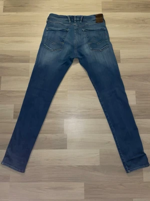 Blå slim fit replay Anbass jeans - Säljer ett par blå slim fit jeans från replay med klassisk femficksdesign och snygga slitningar framtill. Jeansen har normal midja, bälteshällor och en patch med logga bak i midjan. Perfekta för dig som gillar en smalare passform och tidlös denimstil.