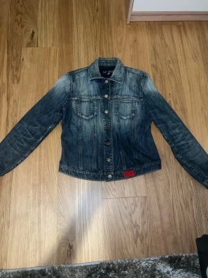 Blå jeansjacka från Armani Jeans - Snygg blå jeansjacka från Armani Jeans med klassisk krage, två bröstfickor med lock och knappar framtill. Jackan har slitna detaljer och en liten röd patch vid nederkanten. Perfekt för en cool och avslappnad stil.