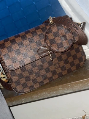 Louis Vuitton brun axelremsväska - Snygg brun axelremsväska från Louis Vuitton i klassiskt rutigt Damier-mönster. Väskan har justerbar axelrem med logotyp, guldiga metalldetaljer och en matchande rund coin purse. Tillverkad i canvas och läder.