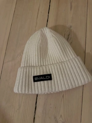 Vit ribbad mössa från Bvaldi - Snygg vit ribbstickad mössa från Bvaldi med uppvikt kant och svart logotypmärke framtill. Tillverkad i en mjuk mix av viskos, nylon och polyester som ger en skön stretchig passform. Perfekt för kalla dagar och enkel att matcha med olika outfits.