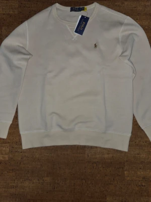 Caramel sweatshirt från Polo Ralph Lauren - Klassisk vit sweatshirt från Polo Ralph Lauren med rund halsringning och broderad logga på bröstet. Tröjan har ribbade muddar vid ärmslut och nederkant samt en enkel, stilren design. Perfekt för en clean och avslappnad look.