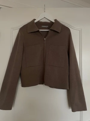 Brun ziptröja från Åhléns - Snygg brun ziptröja från Åhléns med krage och två stora fickor framtill. Tröjan är stickad i ett mjukt material och har en rak, något boxig passform. Perfekt att slänga på sig över en t-shirt för en chill look.