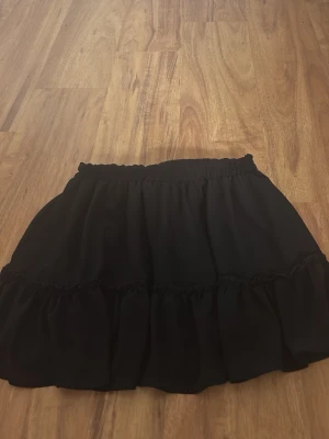 Svart volangkjol  - Säljer en svart kortkjol  med bred resår i midjan och dubbla volanglager. Kjolen har en luftig och flowy känsla, perfekt för att styla med en crop top eller hoodie. Enkel att matcha och superbekväm. Rätt liten i storlek 