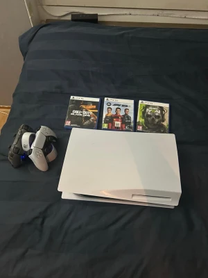 PlayStation 5 disk edition - Säljer en PlayStation 5 i vit färg med två handkontroller, en i svart camo och en i vitt. Konsolen har en futuristisk design och är tillverkad i plast. Tre PS5-spel medföljer: Call of Duty, F1 22 och Halo. Perfekt för gamingkvällar och multiplayer. Det kommer även med en Sony DualSense Charging Station.