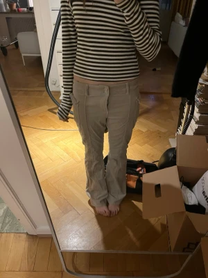 Beigea manschester cargobyxor lågmidjade bootcut  - Säljer ett par beige cargobyxor med bootcut och stora sidofickor. Byxorna har dubbla knappar i midjan och synliga sömmar längs benen för en cool look. Perfekta för dig som gillar avslappnad stil och praktiska detaljer.