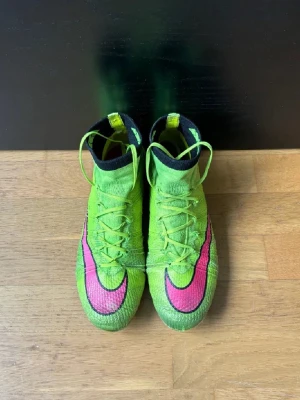 Nike Mercurial Superfly 4 Elite - Säljer ett par Nike Mercurial Superfly fotbollsskor i neon-gult med rosa och svart detaljer. Skorna har hög krage och är tillverkade i syntetmaterial med stickad ovandel för extra komfort och passform. Perfekta för snabba spelare som vill ha grymt grepp på planen.