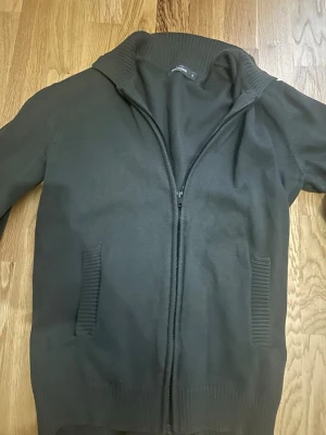 Mörkgrå stickad ziptröja - Säljer en mörkgrå stickad ziptröja med hög krage och ribbade detaljer vid fickor, ärmslut och nederkant. Tröjan har dragkedja framtill och två fickor. Perfekt för lager på lager och enkel att matcha med olika outfits.