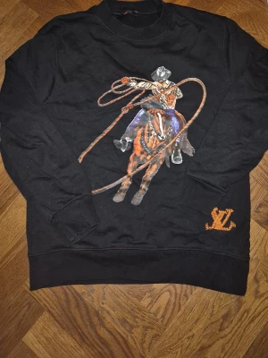 L v sweater - Cool svart sweatshirt med stort tryck av en cowboy på häst som svingar ett lasso, både fram och bak. Orange broderad detalj med bokstäverna 'SL' vid nederkanten. Perfekt för dig som gillar unika och statement-plagg.