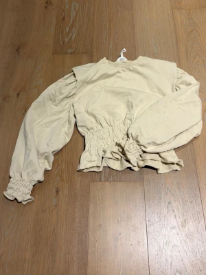 Beige blus med puffärmar från OBJECT - Säljer en beige blus från OBJECT med puffiga långärmar och smockad midja samt ärmslut. Blusen har en rund halsringning och är tillverkad i mjuk bomull. Perfekt för dig som gillar trendiga detaljer och en relaxed vibe.