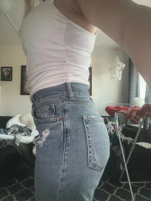 Blå high waist mom jeans - Säljer ett par blå jeans med slitningar på framsidan. Jeansen har klassisk femficksdesign, bälteshällor och knappgylf. Perfekta för en avslappnad och trendig look.