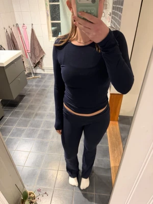 Mörkblå loungewear-set med långärmad topp - Säljer ett mörkblått loungewear-set med långärmad, figurnära topp och matchande byxor med bootcut passform. Perfekt för att chilla hemma och hålla sig bekväm. Materialet är mjukt och stretchigt, vilket gör det extra skönt att bära.