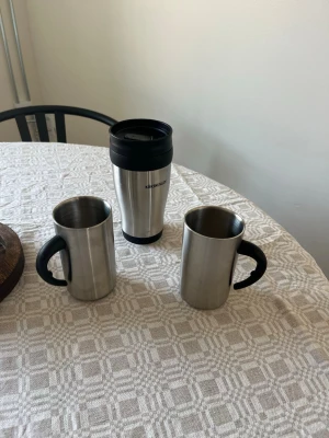 Rostfria muggar och termosmugg - Säljer två rostfria muggar med svarta handtag och en termosmugg från KÖKSMÄSTARE. Perfekt för kaffe eller te på språng. Muggarna har enkel design i metall och termosmuggen har svart lock och greppvänlig form.