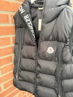 Svart dunväst från Moncler med huva - Snygg svart dunväst från Moncler med huva och två dragkedjefickor framtill. Västen har Moncler-logga på bröstet och broderad text på insidan av kragen. Perfekt för lager-på-lager och streetstyle. Passar även M