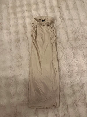 Beige långkjol med rynk - Trendig beige långkjol med rynkade detaljer längs sidorna och hög krage. Perfekt för dig som gillar minimalistisk och stilren design. Materialet känns mjukt och stretchigt, troligen polyester eller bomull.