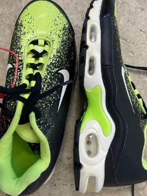 Nike Air Max Plus svart/neongul - Nike Air Max Plus sneakers med svart och neongul färg, vit sula och synliga luftbubblor. Skorna har snörning och den klassiska Swoosh-loggan på sidan. Ovandelen har ett coolt stänkmönster i neongult och svart. Perfekta för dig som vill sticka ut.