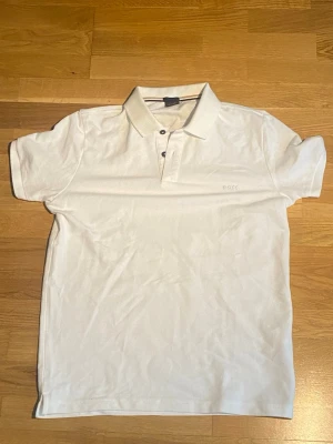 Vit pikétröja från BOSS - Snygg vit pikétröja från BOSS med klassisk krage och tre knappar framtill. Regular fit-modell i mjukt bomullsmaterial. Diskret BOSS-logga broderad på bröstet. Perfekt till jeans eller shorts för en clean och stilren look.