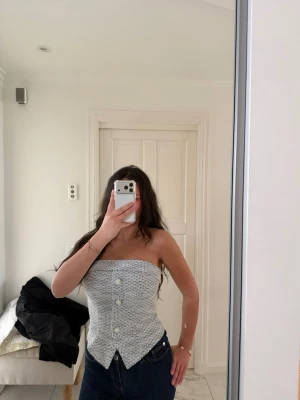 Grå stickad tubtopp med knappar - Snygg grå stickad tubtopp med dekorativa knappar framtill och en unik spetsig nederkant. Toppen är ärmlös och har en croppad passform som ger en trendig look. Perfekt att styla med jeans eller kjol för en cool outfit.