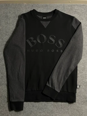 BOSS Hugo Boss – Black & Grey Sweatshirt (S) - Modern and clean BOSS Hugo Boss sweatshirt in a stylish black–grey colorway. Size S, length 64 cm, width 45 cm – a great fit for a slim, contemporary silhouette. The front features the embossed BOSS logo, giving the piece a premium and understated look.  ✅ Condition: Excellent – no wear, no stains 💯 Authenticity: 100% genuine, original Hugo Boss branding 📦 Shipping: Sent immediately after purchase – fast and smooth delivery  This sweatshirt blends comfort, quality, and minimalist design, making 