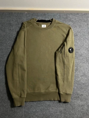 C.P. Company sweatshirt - Authentic C.P. Company sweatshirt in a stylish olive green tone. Size S, length 66 cm, width 51 cm – fits perfectly for a modern, relaxed look. The sleeve features the signature lens detail, a hallmark of C.P. Company’s technical design.  ✅ Condition: Excellent overall – note a small, barely noticeable hole near the lens, visible in the photos but discreet in person 💯 Authenticity: 100% genuine, easily verified on certilogo.com 📦 Shipping: Sent immediately after purchase