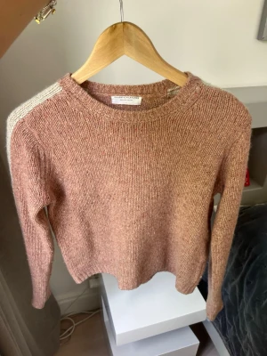 Rosa stickad tröja med vita detaljer - Mysig stickad tröja från Filippa K i en mjuk rosa nyans med vita ränder längs axlar och ärmar. Tröjan har rund halsringning, långa ärmar och är tillverkad i en ullblandning som känns både varm och skön. Perfekt för lager på lager under kyliga dagar.