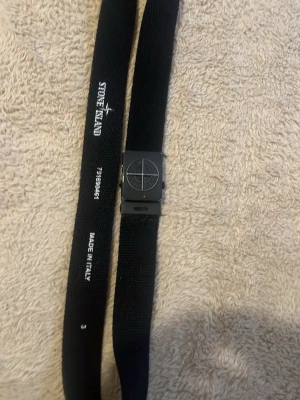Svart bälte från Stone Island - Snyggt svart bälte från Stone Island med metallspänne och logga på spännet. Tillverkat i Italien och har en stilren, enkel design som passar till de flesta outfits. 