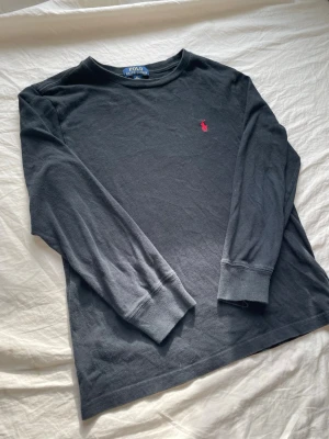 Svart långärmad tröja Polo Ralph Lauren - Svart långärmad tröja från Polo Ralph Lauren med klassisk rund halsringning. Tröjan har en liten röd broderad logga på bröstet och är gjord i mjuk bomull. Perfekt för en stilren och enkel look.