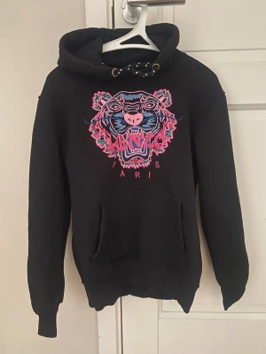 Svart Kenzo hoodie med tigertryck - Svart hoodie från Kenzo med ikoniskt färgstarkt tigertryck på bröstet och texten 'Paris'. Hoodien har huva med snörning och en stor magficka. Materialet är mjukt och bekvämt, perfekt för en cool och avslappnad stil. Stl M men liten i stl , skulle säga S . 
