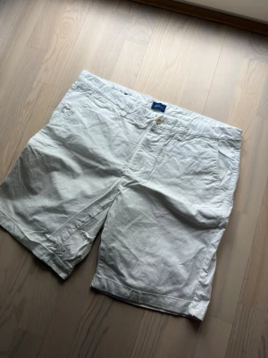 Vita eleganta shorts från Gant - Säljer ett par klassiska vita shorts från Gant i linneblandning. De har raka ben, fickor fram och bak samt stängs med knapp och dragkedja. Perfekta för sommaren och ger en clean och fräsch look.
