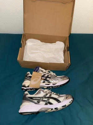 ASICS Helt Nya Silver Cream - Snygga ASICS sneakers i vitt och silver med svarta detaljer. Skorna har en sportig design med mesh- och syntetmaterial, samt en chunky sula med svartvitt mönster. Perfekta för dig som gillar streetstyle och bekväma skor med cool look. Helt Nya, Storlek 44