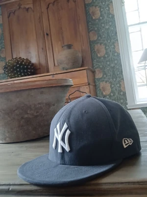 Mörkgrå New Era NY keps - Snygg mörkblå keps från New Era med klassiska New York Yankees-loggan broderad i vitt framtill. Kepsen har rak skärm och broderad New Era-logga på sidan. Perfekt för dig som gillar streetstyle och sportiga accessoarer. Storlek 7 1/8