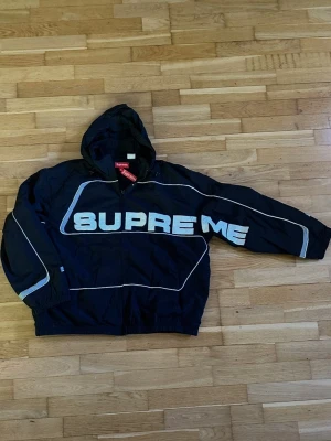 Svart Supreme vindjacka med huva - Svart vindjacka från Supreme med stor vit logga över bröstet och ärmen. Jackan har huva, dragkedja och vita kontrastlinjer längs ärmar och kropp. Perfekt för streetwear-stil och snygg att bära till jeans eller joggers.