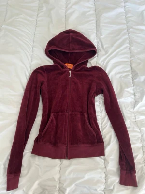 Juicy Couture vinröd velour hoodie - Juicy Couture hoodie i vinröd velour med dragkedja framtill, huva och kängurufickor. Baksidan har broderi med texten 'Love Luxury Juicy Couture' och en krona. Perfekt för en chill och trendig look.