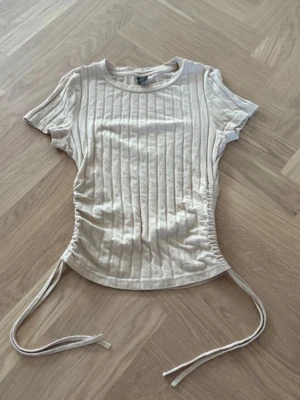 Beige ribbad topp med snörning Shein - Trendig beige ribbad topp från Shein med korta ärmar och snörning i sidorna. Toppen har en rund halsringning och är tillverkad i ett mjukt material som sitter snyggt på kroppen. Perfekt för en stilren och avslappnad look.