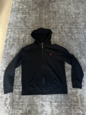 Svart hoodie från Polo Ralph Lauren - Svart hoodie med dragkedja från Polo Ralph Lauren. Klassisk design med huva, känguruficka och röd broderad logga på bröstet. Perfekt för en avslappnad och stilren look.