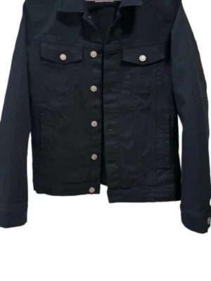 Svart jeansjacka jack and jones - Stilren svart jeansjacka med klassisk krage och silverfärgade knappar framtill. Två bröstfickor med lock och knapp, samt raka ärmar med knapp vid ärmslut. 