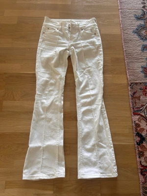 Vita utsvängda jeans med medel låg midja - Säljer ett par vita jeans med medel låg midja och utsvängda ben. Jeansen har dubbla knappar i midjan, klassiska fem fickor och markerade sömmar längs benen. Perfekta för en trendig och fräsch look.