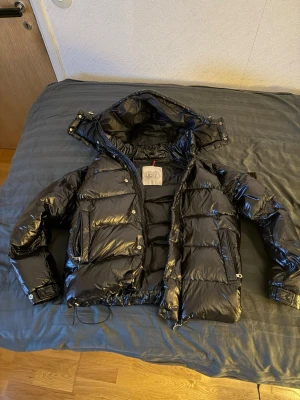 Moncler jacka - Säljer en svart glansig dunjacka från Moncler med avtagbar huva och dragkedja framtill. Jackan har klassisk puffer-design, tryckknappar och innerfoder med Monclers ikoniska serietidningsmärke. NFC-Scan fungerar perfekt 