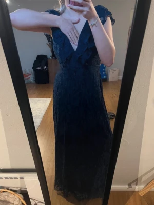 Marinblå långklänning med spets - Elegant marinblå långklänning med v-ringning och volangdetaljer framtill. Klänningen har spetsmaterial, korta ärmar och en öppen rygg som ger en snygg och trendig look. Perfekt för bal eller annat!💞